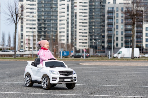 Klein meisje rijdt in een grote witte elektrische speelgoedauto voor kinderen op de parkeerplaats bij het woonhuis