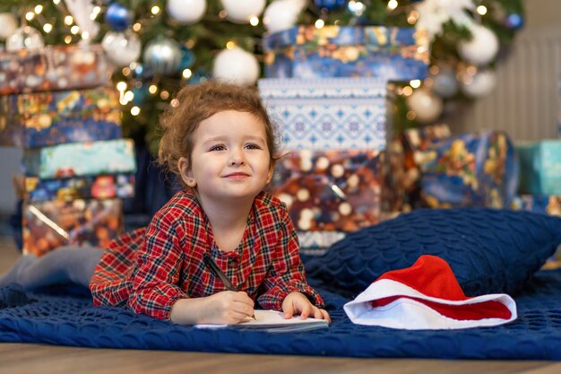 Foto klein meisje schrijft brief aan de kerstman. kinderdroom bij kerstboom over wens, cadeau op nieuwjaar