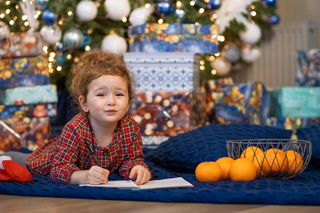 Foto klein meisje schrijft brief aan de kerstman. kinderdroom bij kerstboom over wens, cadeau op nieuwjaar