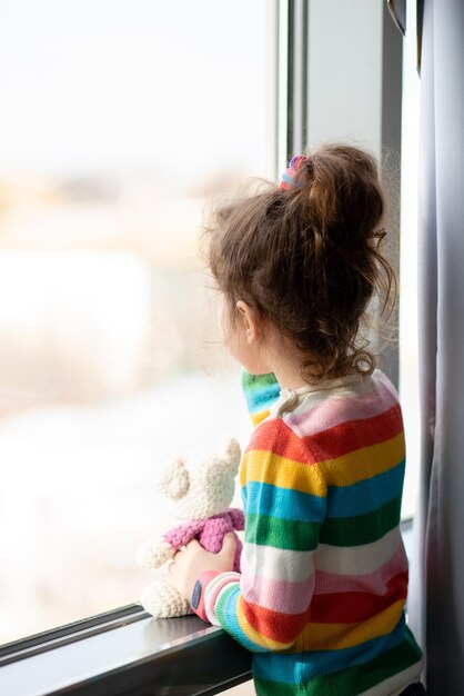 Klein meisje staat bij een raam met teddybeer in haar handen Ze is gekleed in een gekleurde trui