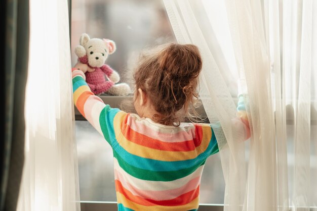 Klein meisje staat bij een raam met teddybeer in haar handen