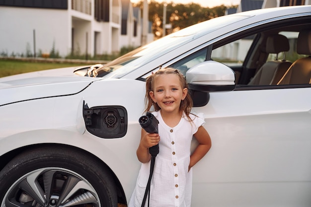 Klein meisje staat in de buurt van elektrische auto met oplaadkabel in handen