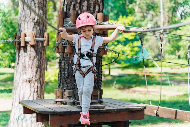 Klein meisje staat op een touw en houdt een touw met zijn handen vast. Een kind in een touwpark passeert obstakels