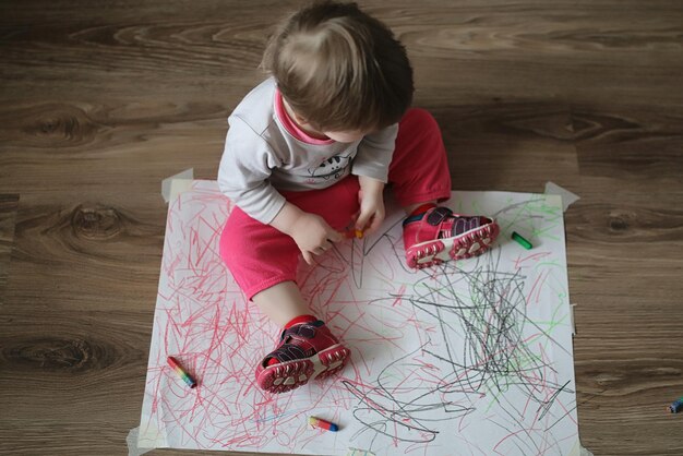 Klein meisje tekent met kleurpotloden op een groot wit vel papier, liggend op de vloer. Het kind is aan het schilderen op de vloer.