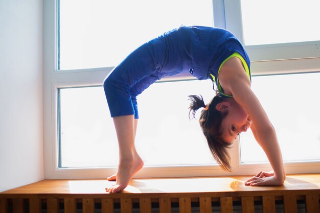 Klein meisje training thuis in bridge pose. Schattige jongen traint op een houten vensterbank binnen. Kleine donkerharige vrouwelijk model in sportkleding heeft oefeningen bij het raam in haar kamer.