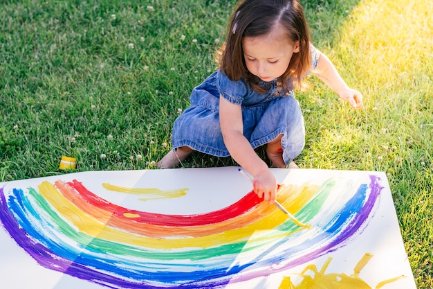 Klein meisje van 2-4 jaar schildert regenboog en zon op groot vel papier, zittend op groen gazon in zonlicht