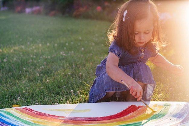 Foto klein meisje van 2-4 jaar schildert regenboog en zon op groot vel papier, zittend op groen gazon in zonlicht