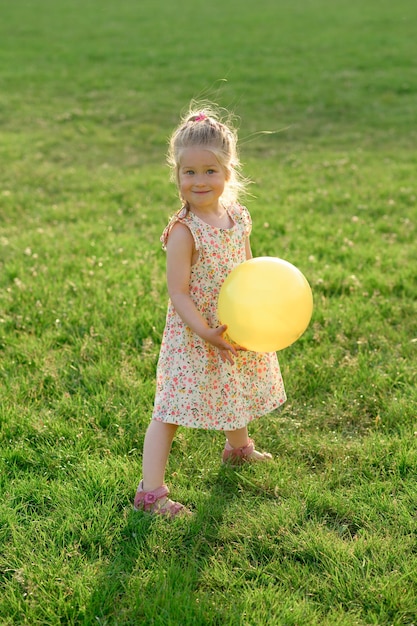 Klein meisje van 3 jaar oud in een zomerjurk op een open plek met een gele bal in haar handen Het meisje is gelukkig