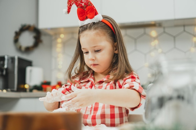 Klein meisje van 3 jaar oud in rode kerstmuts en geruit hemd bereidt deeg voor peperkoekkoekjes in de keuken