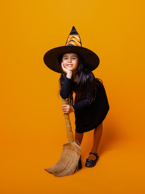 Klein meisje verkleed als Halloween-heks in een zwarte jurk en hoed vliegt op een bezemsteel op een gele achtergrond.