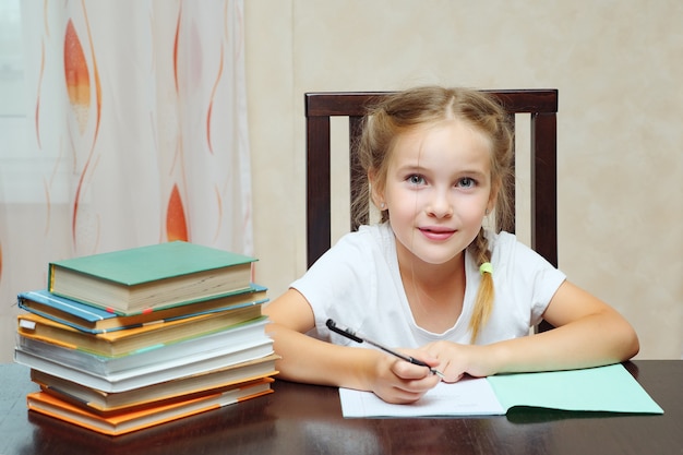 Klein meisje zit aan tafel met stapel boeken en doet huiswerk en kijkt naar de camera
