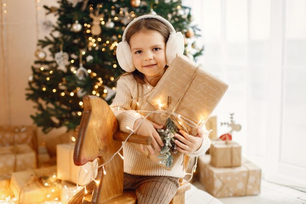 Klein meisje zit in de buurt van de kerstboom op een houten hobbelpaard speelgoed