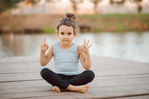 Foto klein meisje zit in de natuur in de buurt van rivier of meer en beoefent yoga gezonde levensstijl