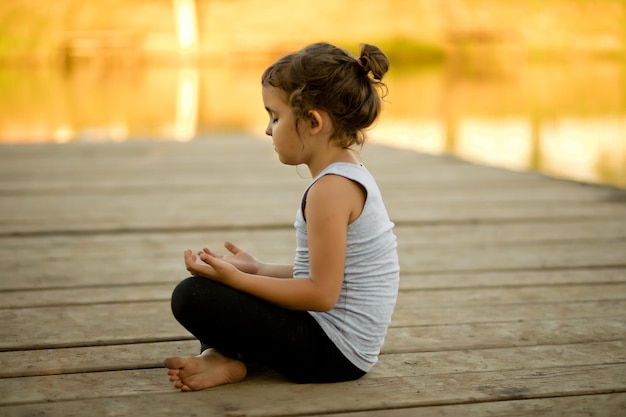 Klein meisje zit in de natuur in de buurt van rivier of meer en beoefent yoga gezonde levensstijl