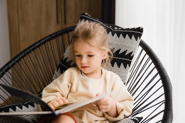 Klein meisje zit in een stoel bij het raam en leest een boek