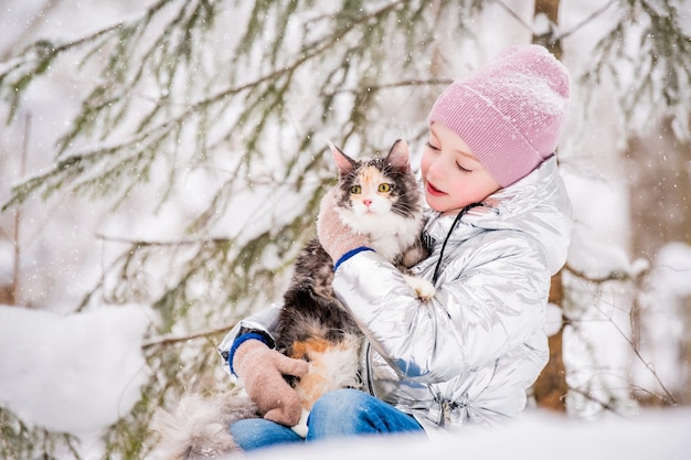 Klein meisje zit met een kat in een omhelzing in de sneeuw in het winterbos