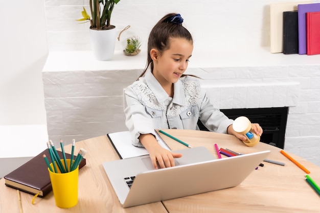 klein meisje zit met een laptop aan tafel en houdt een zandloper vast, wachtend op het einde van de online lessen