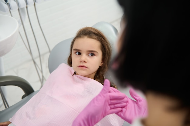 Klein meisje zittend in tandartsstoel, kijkend naar een kinderhygiënist die haar leert over mondhygiëne en preventie van tandbederf tijdens tandheelkundig bezoek in witte, schone, moderne medische kast