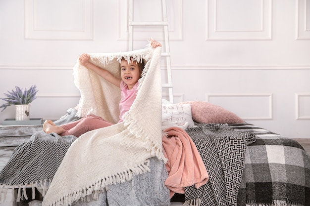 Klein meisje zittend op bed met mooie dekens op een lichte achtergrond van de slaapkamer.