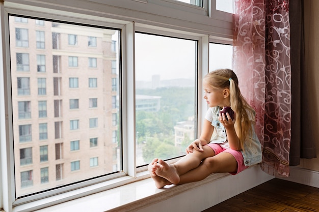 klein meisje zittend op de vensterbank thuis, kijkend in het venster en rode appel te houden