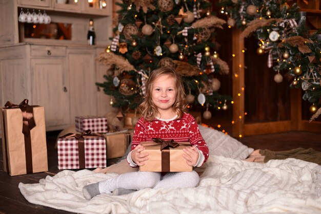 Klein meisje zittend op de vloer in de buurt van de kerstboom en geschenk in haar handen te houden