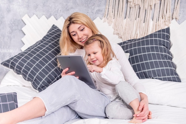 Klein mooi meisje drukt op het scherm van de tablet die met moeder in bed ligt