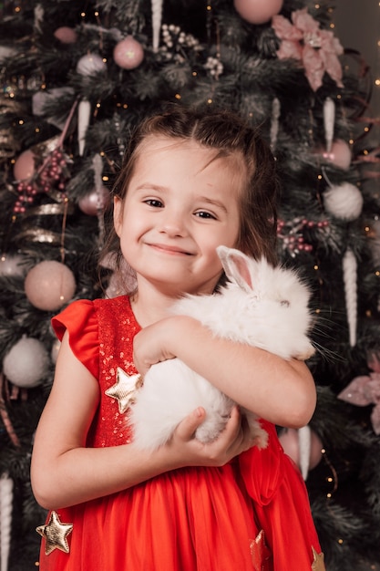 Klein mooi meisje lacht en houdt een wit pluizig konijn in haar armen bij de kerstboom