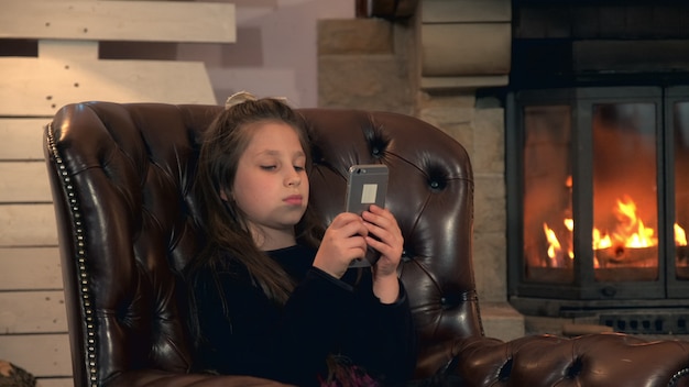 Klein mooi meisje spelen van games op smartphone in de buurt van de open haard