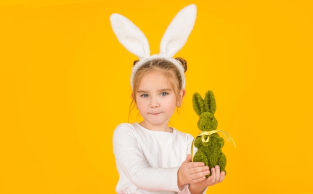 klein paaskind in konijnenoren die speelgoed op gele achtergrond houden.