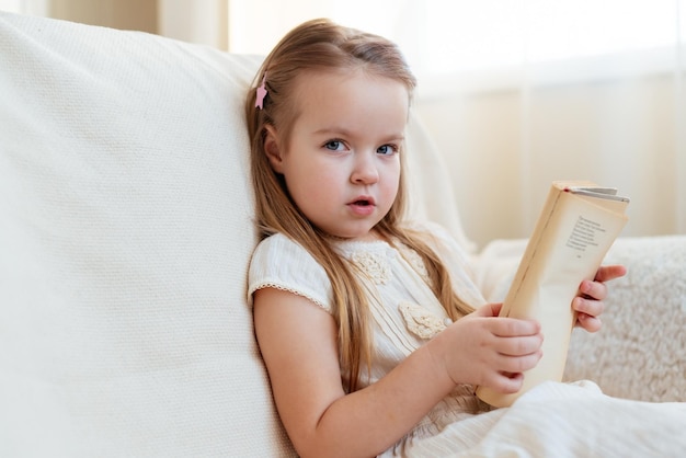 Klein schattig blond meisje leesboek zittend op een bank Kind lezen dromen en verbeeldingsontwikkeling Internationale Kinderboekendag Kinderen houden van lezen Wereldboekendag