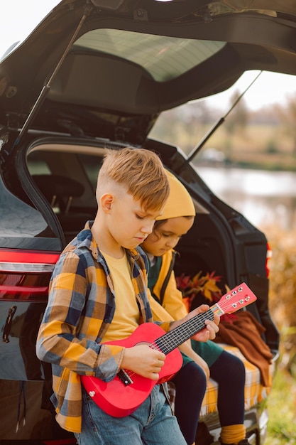 Klein schattig lachend meisje en jongen staan dichtbij op open kofferbak Jongen speelt kleine gitaar Kind rust met haar familie in de natuur herfstseizoen