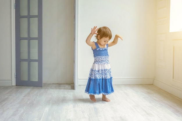 Klein schattig lief lachend meisje in blauwe jurk dansen in fel lichte woonkamer thuis en lachen. Jeugd, kleuterschool, jeugd, ontspannen concept