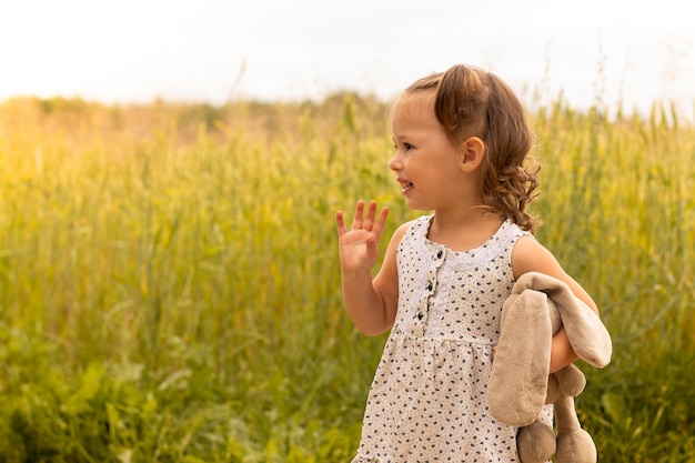 Klein schattig meisje 1-3 met een pluche haas in een lichte jurk die met zijn hand zwaait in een veld met roggeaartjes in de zomer