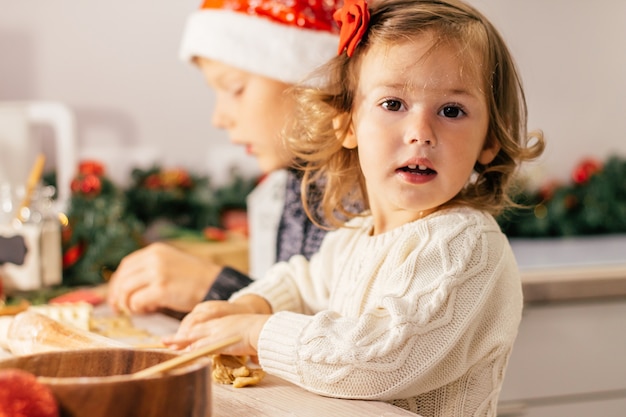 Klein schattig meisje 2-4 met een rode strik en jongen 7-10 in een kerstmuts maken peperkoekkoekjes in de nieuwjaarskeuken.