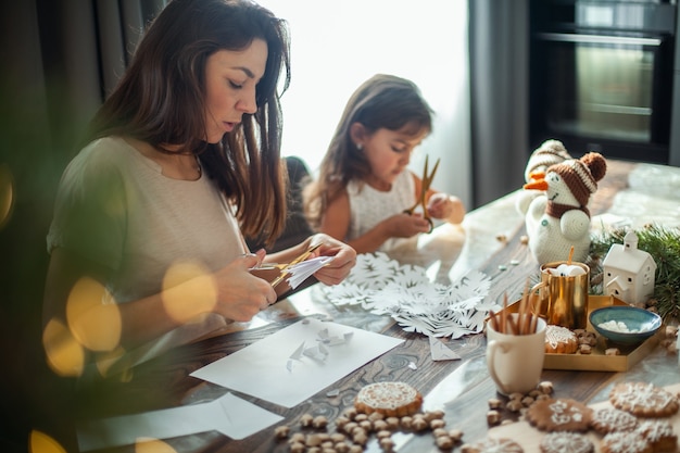 Klein schattig meisje en jonge mooie vrouw snijden sneeuwvlokken van wit papier peperkoek en cacao met...