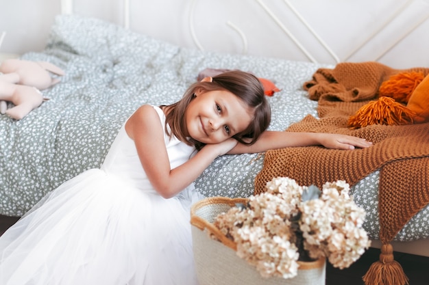 Klein schattig meisje in een mooie witte jurk ontspant in haar lichte slaapkamer