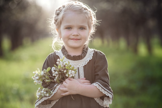 Klein schattig meisje in jurk in bloesemtuin. Schattige baby meisje 3-4 jaar oud bloemen houden over de natuur. Lente portret. Aromatische bloesem en retro vintage concept.