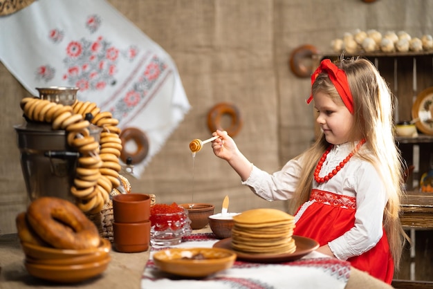 Klein schattig meisje in klederdracht sprinklers pannenkoeken met honing Traditioneel eten voor Shrovedite