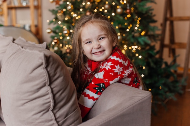 Klein schattig meisje in rode kersttrui speelt thuis bij de kerstboom. Nieuwjaar decoratie.