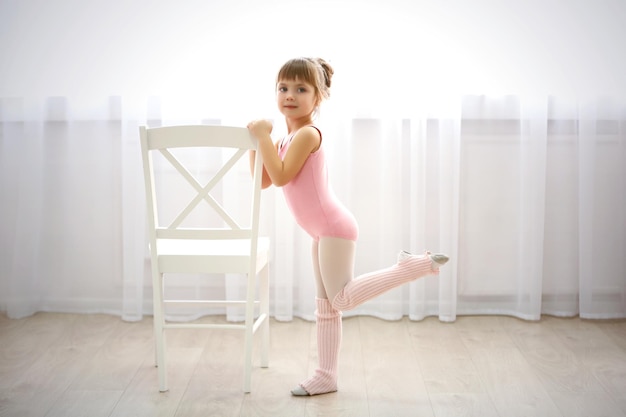 Klein schattig meisje in roze turnpakje dat nieuwe balletbeweging maakt in de buurt van stoel in dansstudio