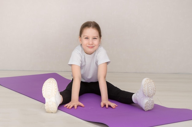 Klein schattig meisje in sportuniform zit op gymnastiekmat met uitgestrekte armen naar voren en glimlacht