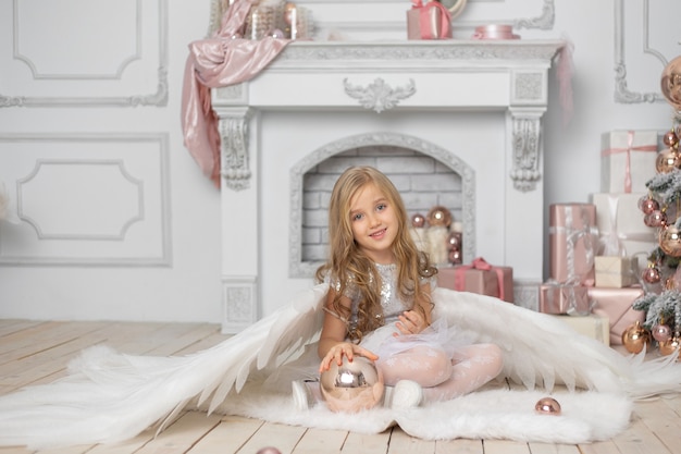 Klein schattig meisje met blond haar met engelenvleugels in de studio naast de kerstboom