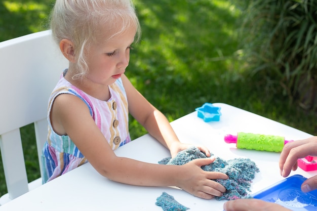 Klein schattig meisje met blond haar spelen met kleurrijk kinetisch zand buiten in de achtertuin in zomerdag. Hobby, vrijetijdsbesteding, knutselen, modelleren en creatief hobbyconcept