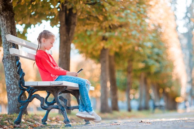 Klein schattig meisje met smartphone in de herfst. jong geitje dat pret heeft bij warme zonnige de herfstdag in openlucht