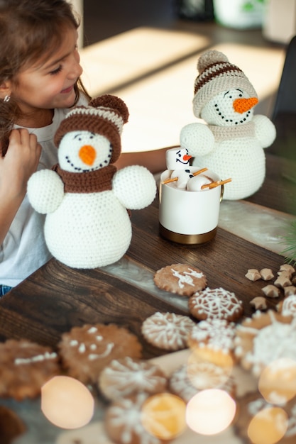 Klein schattig meisje speelt met gebreide sneeuwmannen en eet peperkoek en drinkt cacao met marshmallows
