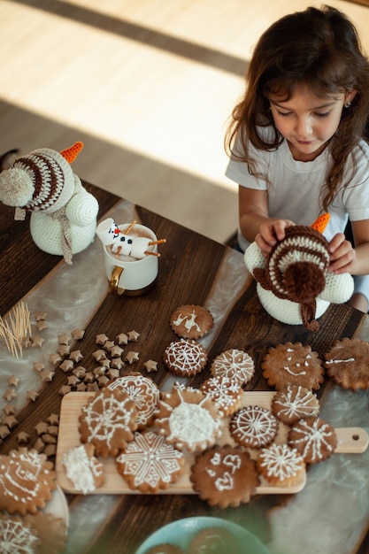 Foto klein schattig meisje speelt met gebreide sneeuwpoppen en eet peperkoek en drinkt cacao met marshmallows.