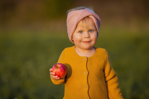 Klein schattig meisje wandelingen in de herfsttuin, houdt een mand met rode appels. portret van een gelukkig meisje in heldere herfstkleren. warme en heldere herfst.