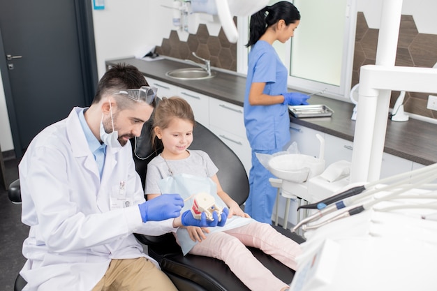 Klein schattig meisje zittend in een stoel en kijken naar valse tanden gehouden door haar tandarts in whitecoat uit te leggen hoe te zorgen voor mondholte