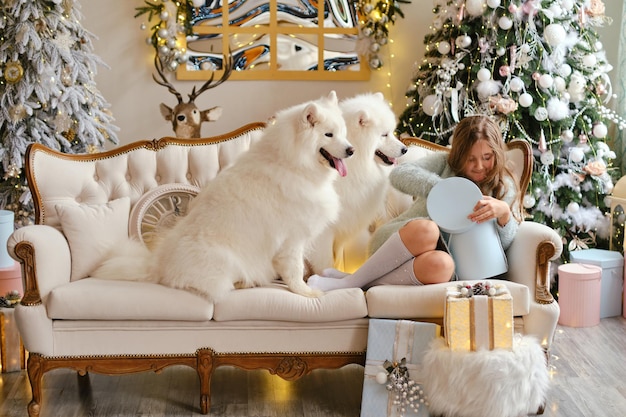 Klein schattig meisje zittend op de bank met twee Samojeed-honden, geschenkdozen openend bij de kerstboom in een prachtig interieur