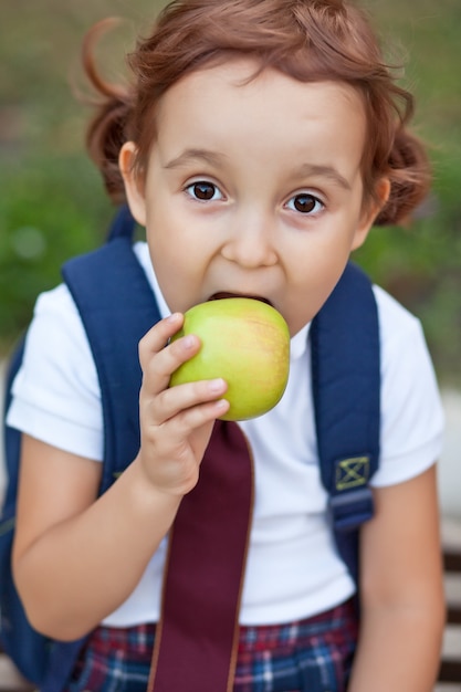 Klein schattig schoolmeisje in uniform zittend op een bankje en een appel etend